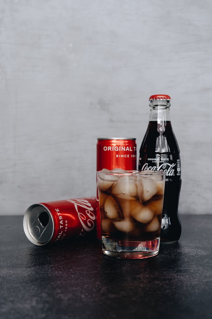 Chilled glass of cola with ice, surrounded by bottles and cans on a dark surface.